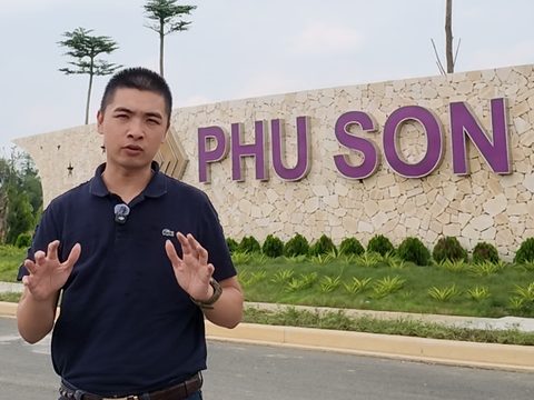 Đức đứng bên đường, nhìn thẳng vào máy, hai tay giơ trước ngực khi đang nói; phía sau là tường đá có dòng chữ PHU SON