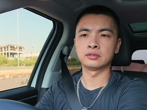 Đức ngồi sau tay lái, nhìn thẳng vào máy; ngoài cửa kính là khối nhà xây thô giữa bãi cỏ ven đường