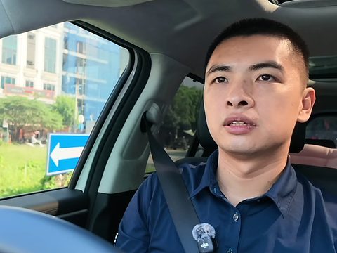 Đức ngồi sau tay lái, mặt hướng về phía máy; ngoài cửa kính là bãi đất trống cỏ mọc um tùm, có biển chỉ hướng màu xanh, xa hơn là dãy nhà